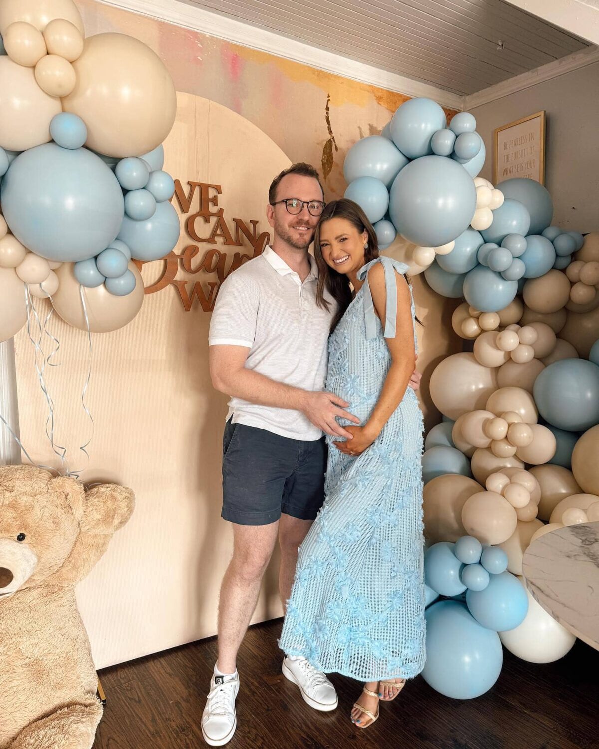 Smiling couple celebrating their baby shower with blue and beige balloon decorations and teddy bear theme.